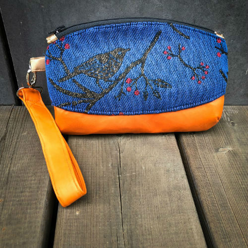A small zippered clutch sewn in orange leather and a babywearing wrap in electric blue, black, and red with a motif of birds in branches.