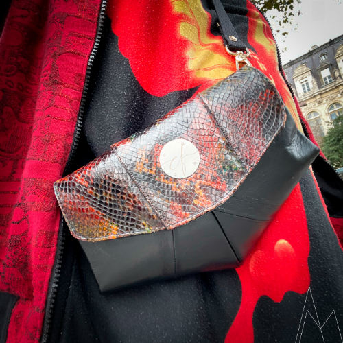 Une petite sacoche pointue avec un rabat rond, fabriquée en cuir d'agneau noir et en cuir de serpent aux tons rouges, portée sur une tunique noir et rouge avec une veste Bombers en tissu d'écharpe de portage noir et rouge.
