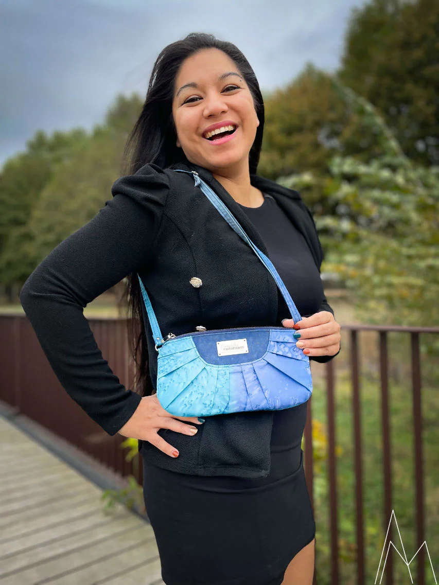 Photo d'une jeune femme à la peau mate portant un sac d'épaule plissé dans les tons de bleus, dans un parc en extérieur, de jour.