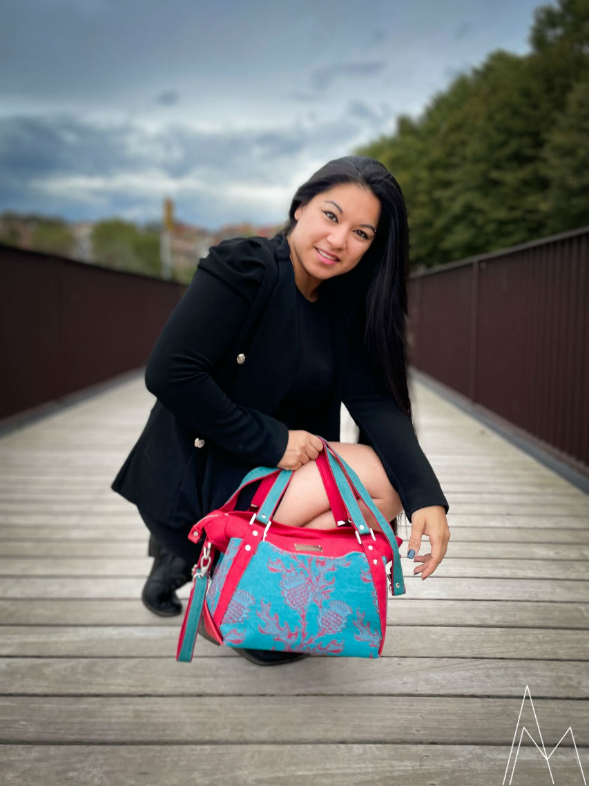 Photo of a young woman with a tan complexion carrying a red and emerald green shoulder bag, in an outdoor park during the day.