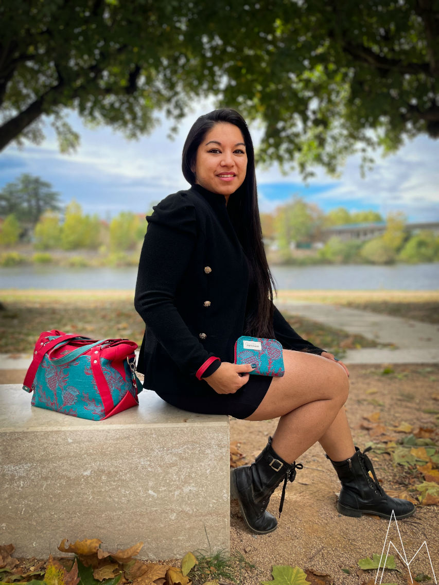 Photo of a young woman with a tan complexion holding a matching red and emerald green wallet next to the shoulder bag laid beside her, in an outdoor park during the day.
