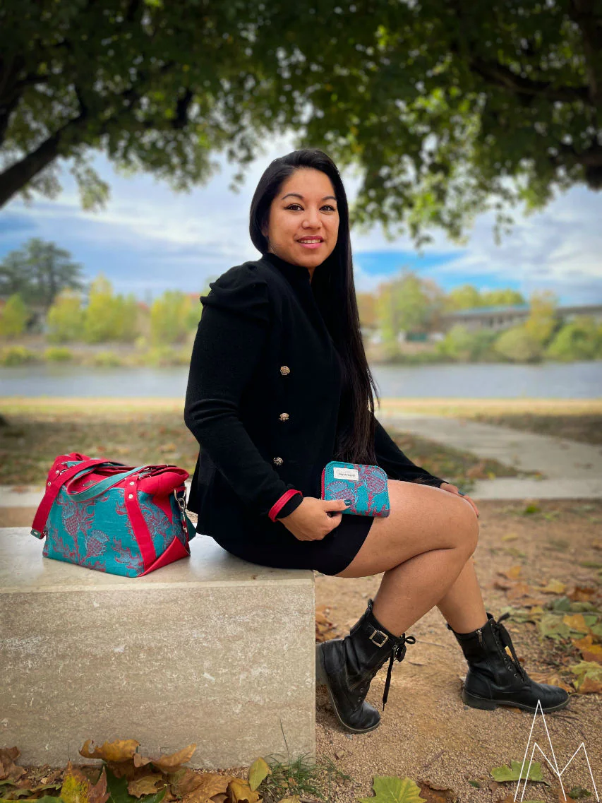 Photo d'une jeune femme à la peau mate portant à la main un portefeuille rouge et vert émeraude assorti au sac d'épaule posé à côté d'elle, dans un parc en extérieur, de jour.