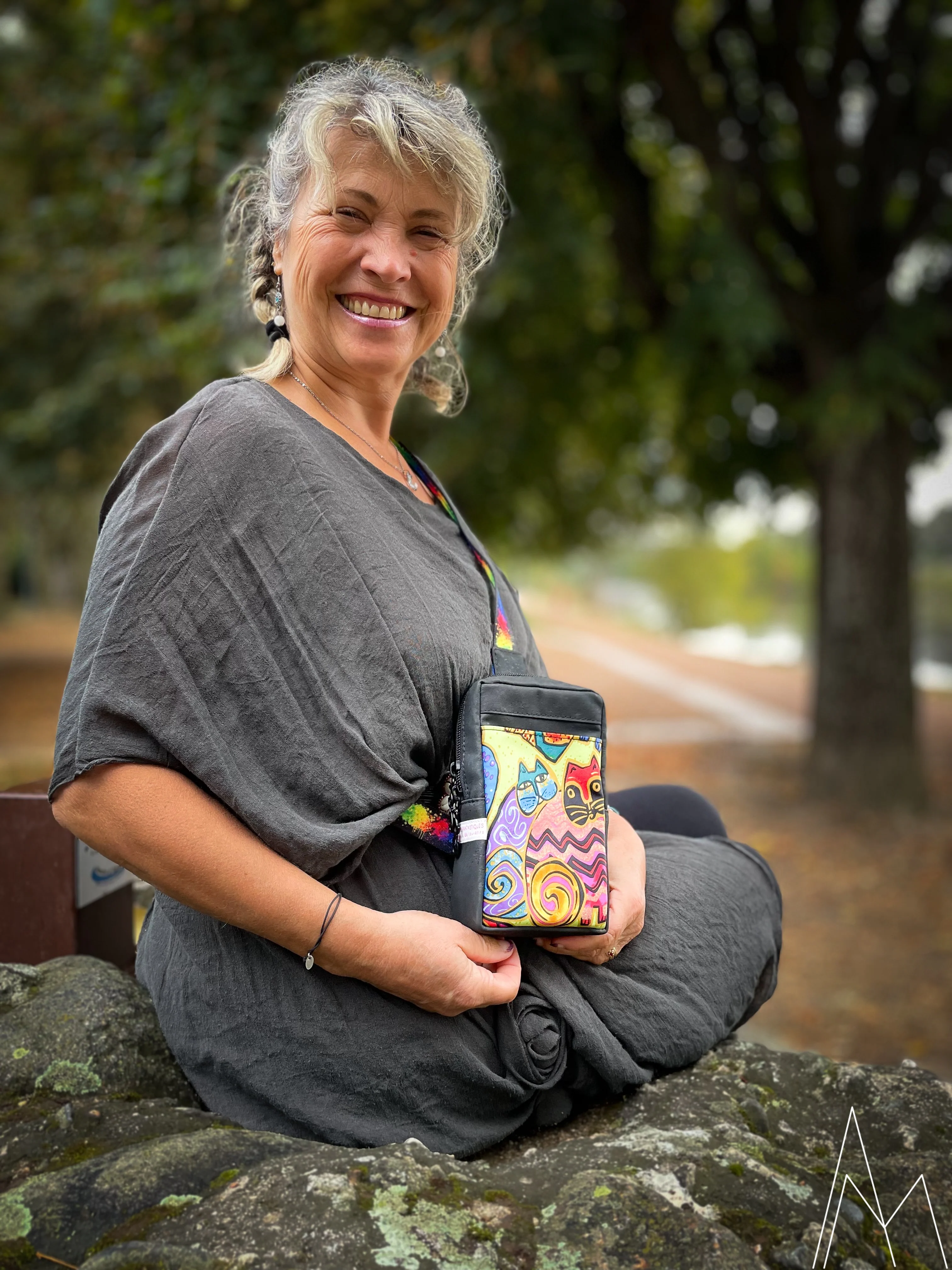 Photo d'une femme mûre portant à la main un sac à dos noir et à motifs de chats bariolés, dans un parc en extérieur, de jour.