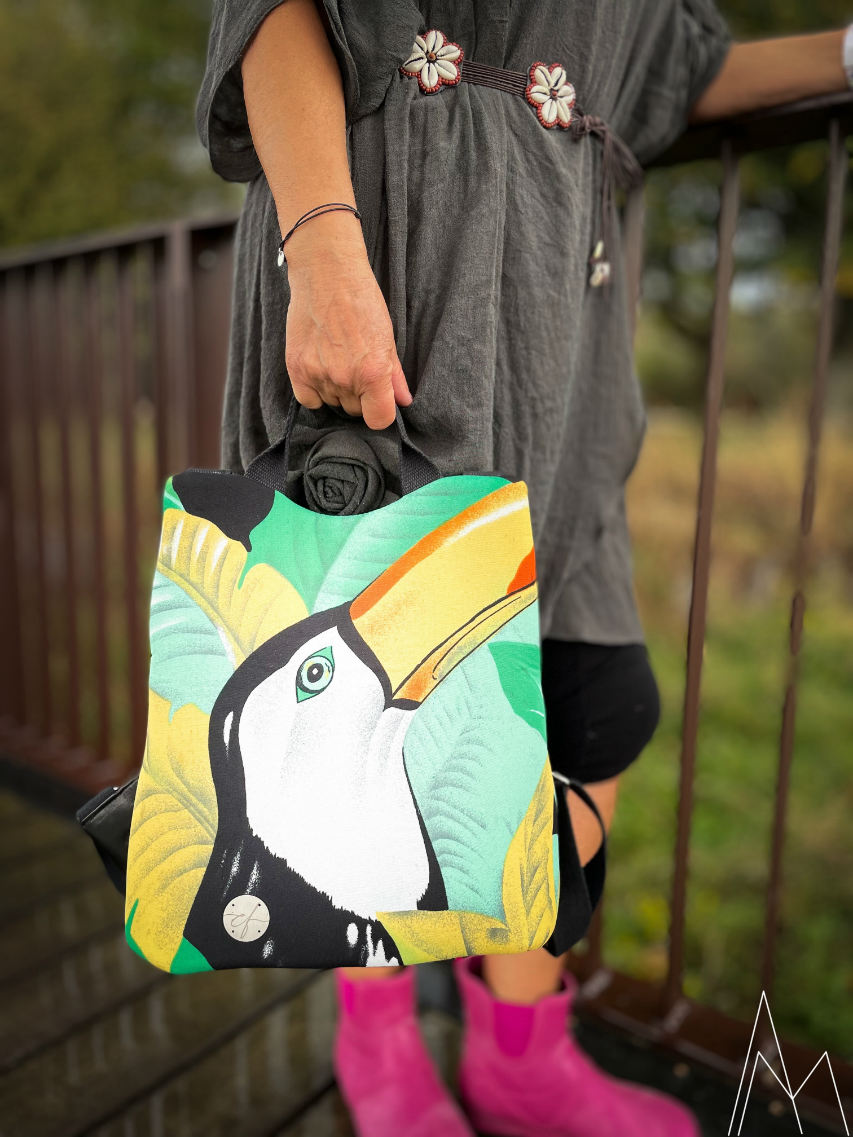 Photo of a mature white woman carrying a backpack with a toucan print by hand, in an outdoor park during the day.