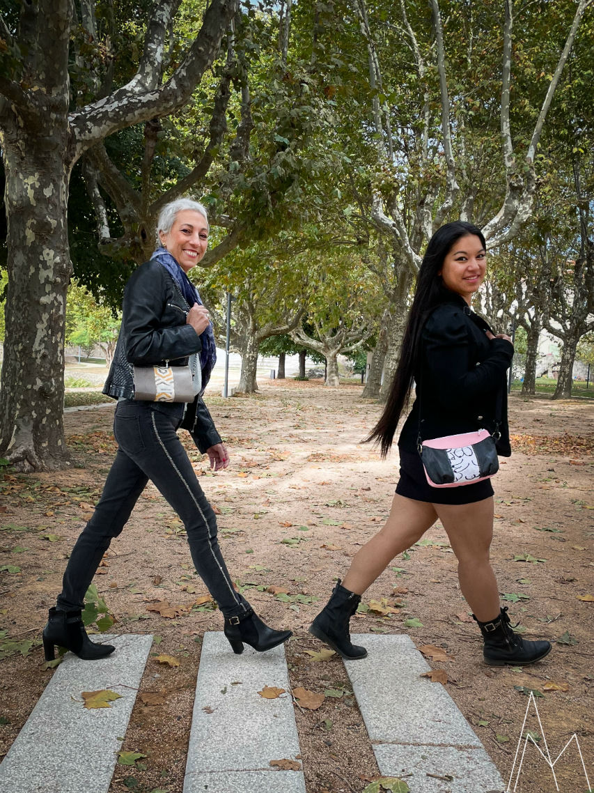 Photo of two women, one young with a tan complexion and one older with fair skin, one carrying a pink and black clutch and the other gray and yellow, in an outdoor park during the day.