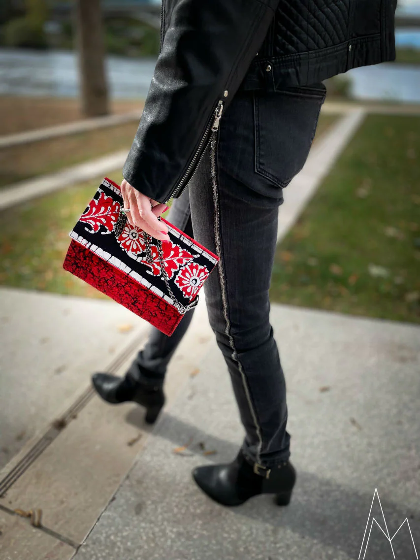 Photo d'une main blanche portant une pochette rouge et noir, dans un parc en extérieur, de jour.