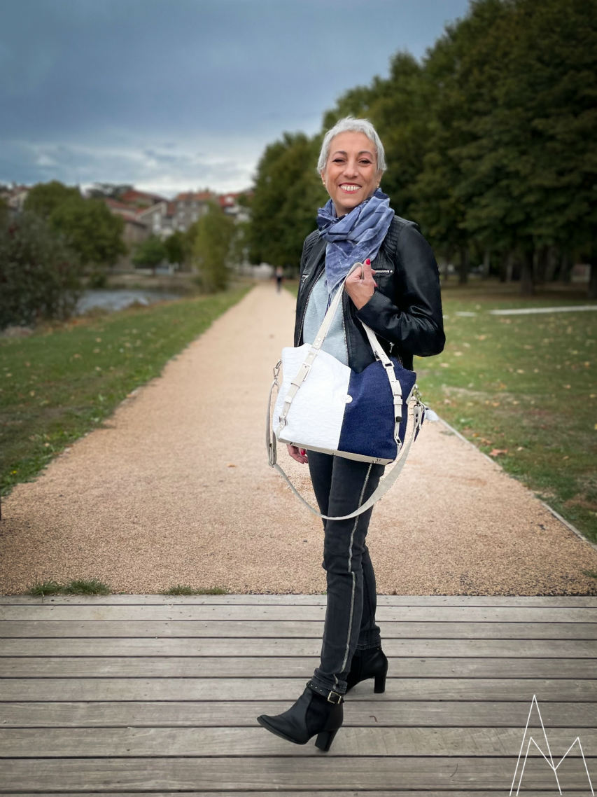 Photo of a mature white woman carrying a navy blue and white shoulder bag, in an outdoor park during the day.