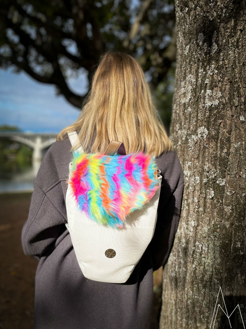Photo of a young blonde woman seen from behind, carrying a white and rainbow backpack, in an outdoor park during the day.