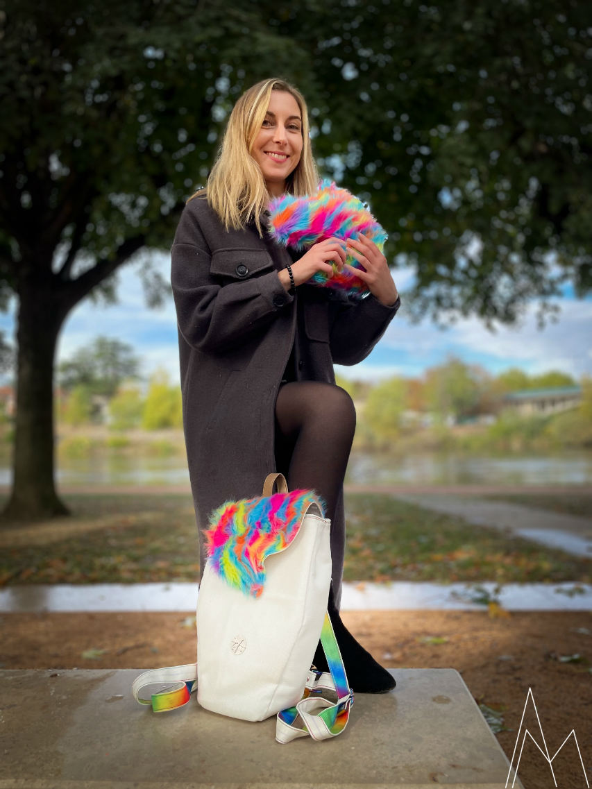 Photo of a young white blonde woman carrying a rainbow faux fur plush and at her feet a white and rainbow backpack, in an outdoor park during the day.