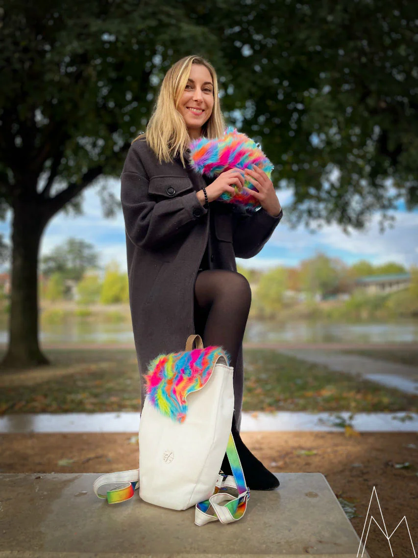 Photo d'une jeune femme blanche et blonde portant une peluche en fourrure synthétique arc-en-ciel et à ses pieds un sac à dos blanc et arc en ciel, dans un parc en extérieur, de jour.