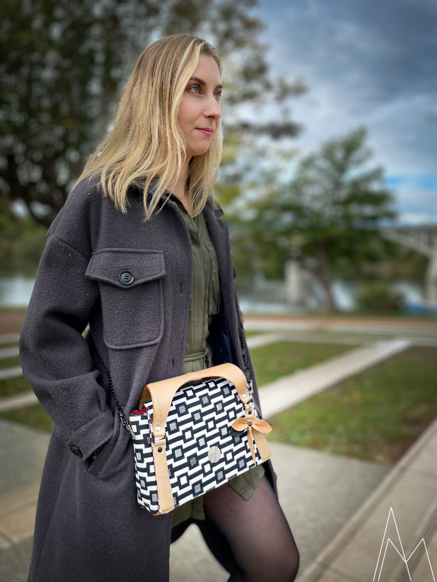Photo of a young white, blonde woman carrying a black and white with caramel shoulder bag, in an outdoor park, during the day.