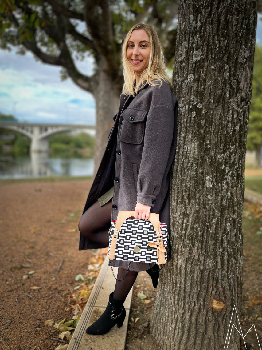 Photo of a young white blonde woman carrying a black and white with caramel shoulder bag, in an outdoor park during the day.