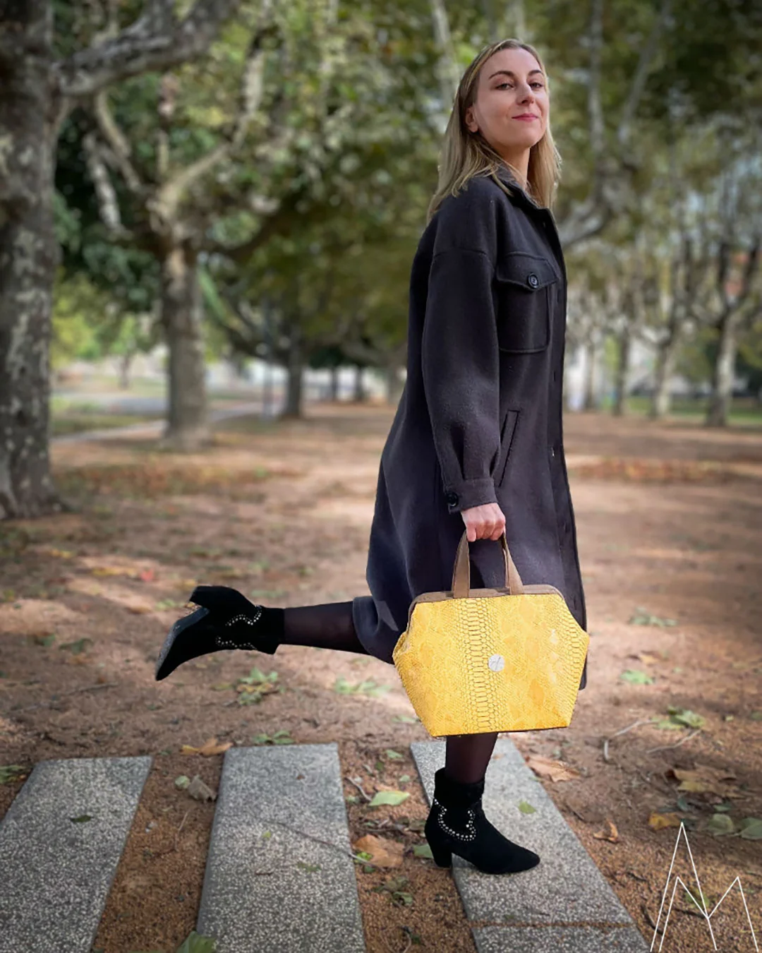 Photo d'une jeune femme blanche et blonde portant un sac à main jaune et marron, dans un parc en extérieur, de jour.