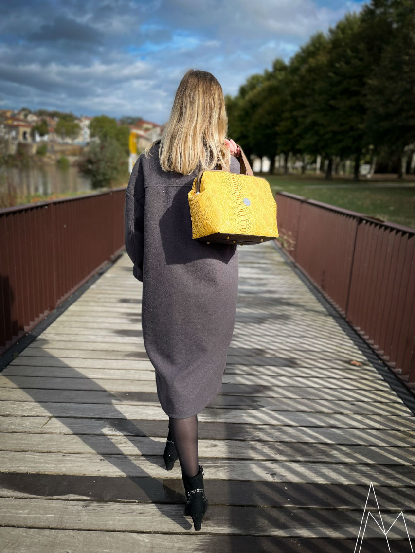 Photo of a young white blonde woman carrying a yellow and brown handbag, in an outdoor park during the day.