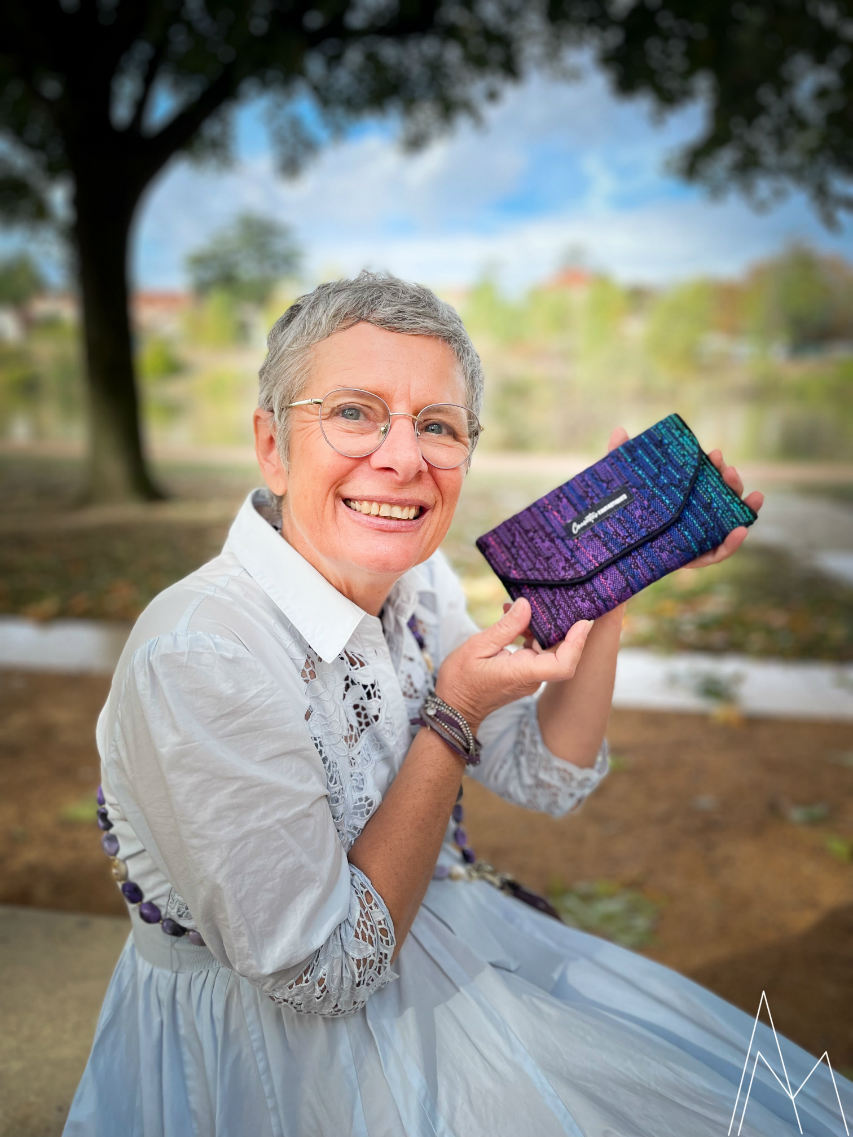 Photo of a mature white woman carrying a wallet in shades of purple and blue, in an outdoor park during the day.