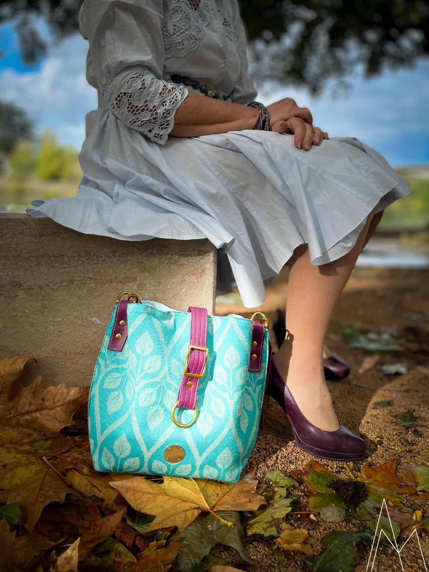 Photo of a mature white woman sitting with a turquoise and plum shoulder bag at her feet, in an outdoor park during the day.