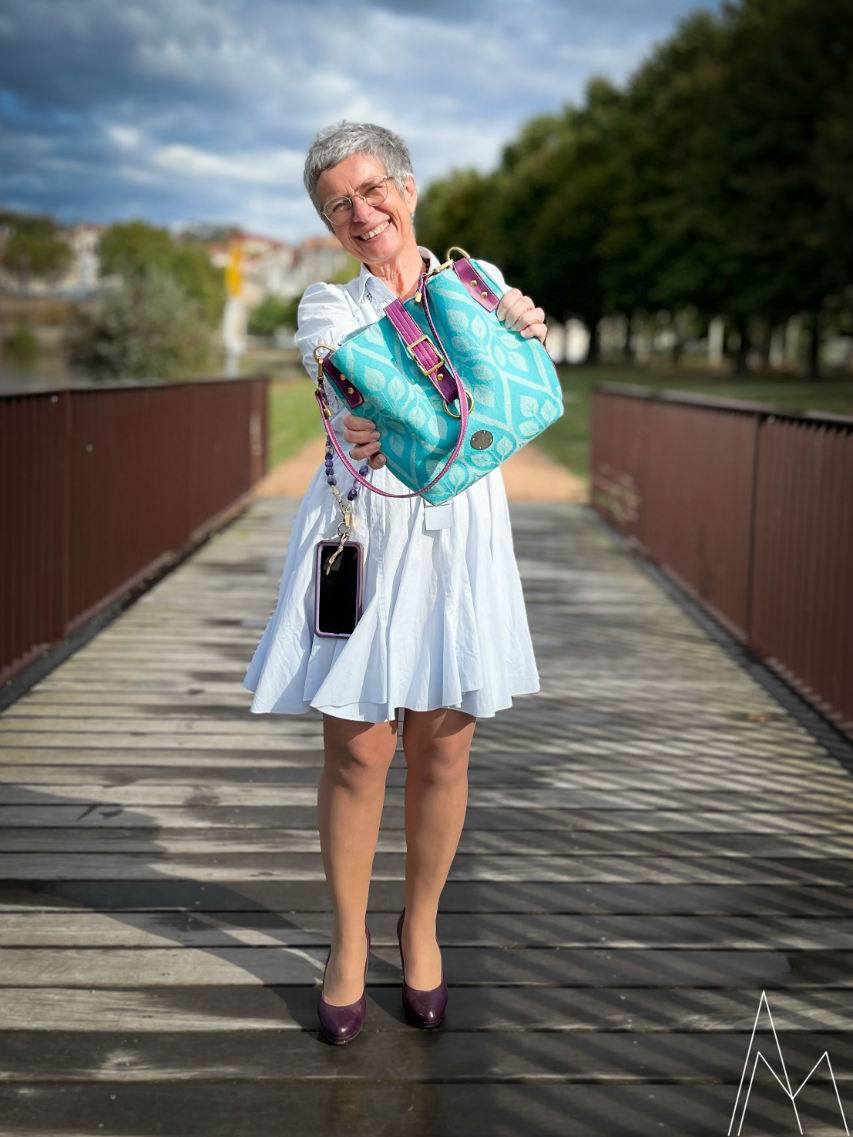 Photo of a mature white woman carrying a turquoise and plum shoulder bag, in an outdoor park, during the day.
