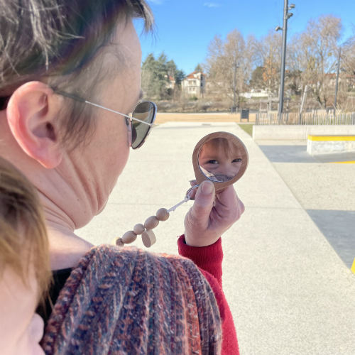 Une femme blanche regarde le reflet de l'enfant blanc porté dans son dos grâce à un miroir en bois