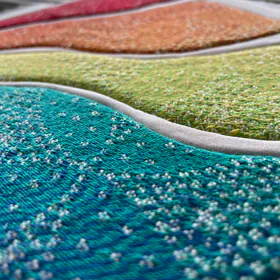Gros plan sur un tableau en kinusaiga fait dans une écharpe de portage arc-en-ciel Oscha : bandes turquoise, verte, jaune, orange et rouge, séparées par de fines rainures blanches, tissage et relief visibles