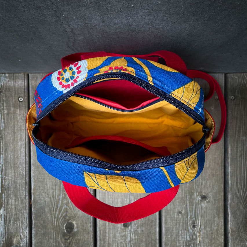 A backpack made from royal blue fabric with yellow, white, and red floral patterns and red embossed fabric on the outside, red and yellow plain linings on the inside.