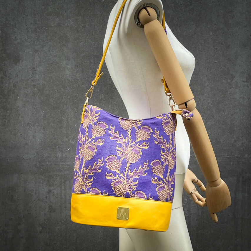 A bucket bag made from babywearing wrap fabric featuring yellow thistles on a purple background with yellow lamb leather.