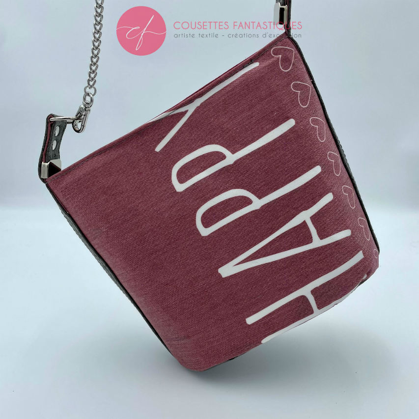 A small “bucket” bag made with delicate pink canvas featuring the word “Happy” and gray ostrich-patterned faux leather.