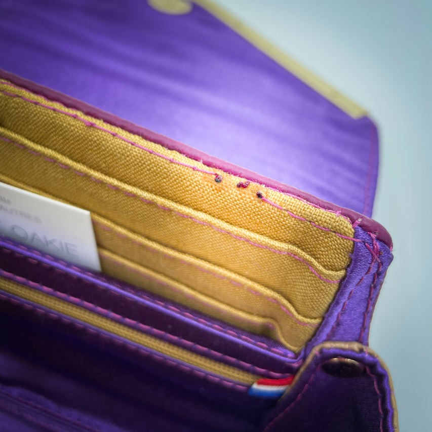 A wallet made from purple and yellow geometrical patterned wrap scrap combined with purple and gold reclaimed faux leathers on the outside, and mustard flax light canvas and deep violet satin on the inside.
