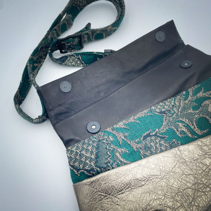 A shoulder bag made from two leathers, gold and black, and a piece of baby wrap fabric in black, green, and gold with a thistle pattern on the outside, and forest green poplin and gold satin on the inside.