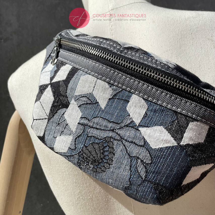 A fanny pack made from babywearing fabric in gray, white, and blue tones with a floral and geometric pattern and gray denim.