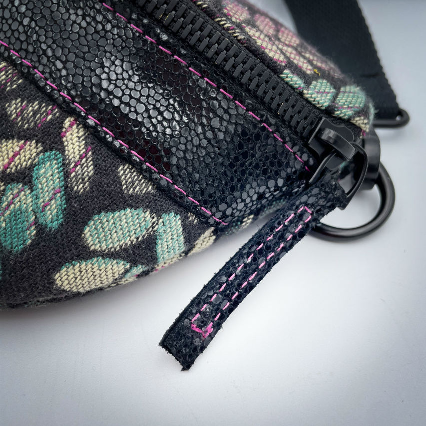 A fanny pack sewn from black patent leather and a scrap of baby wrap fabric with a pattern of green, beige, and pink stitches on a black background, paired with bright pink poplin lining inside.