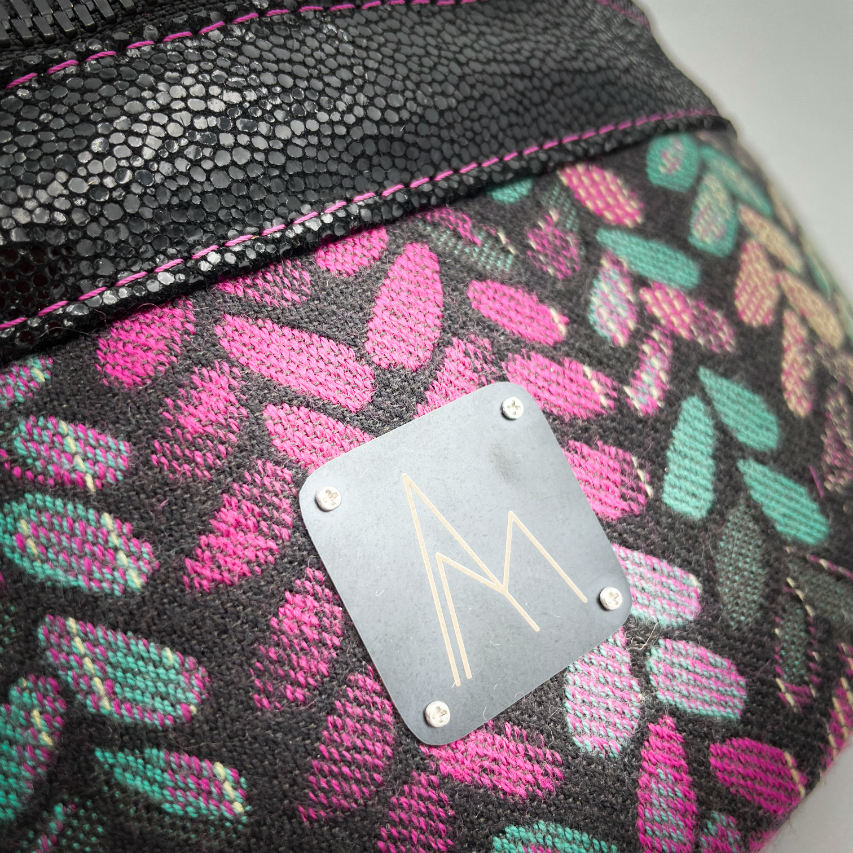 A fanny pack sewn from black patent leather and a scrap of baby wrap fabric with a pattern of green, beige, and pink stitches on a black background, paired with bright pink poplin lining inside.