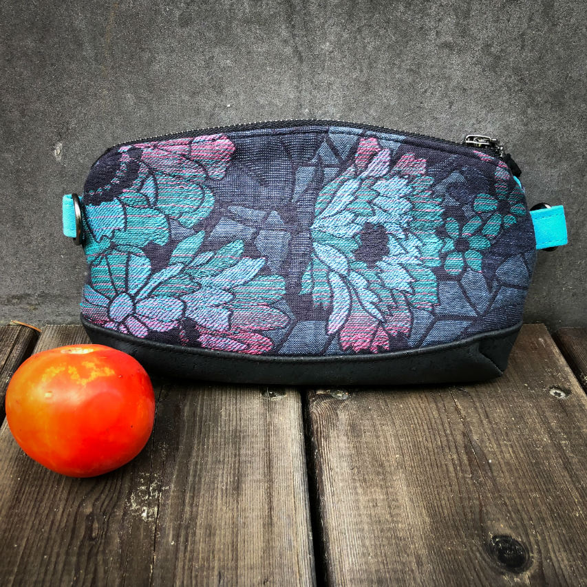 A small zippered clutch sewn in black cork and a babywearing wrap with geometric and floral patterns in shades of blue, green, and pink.