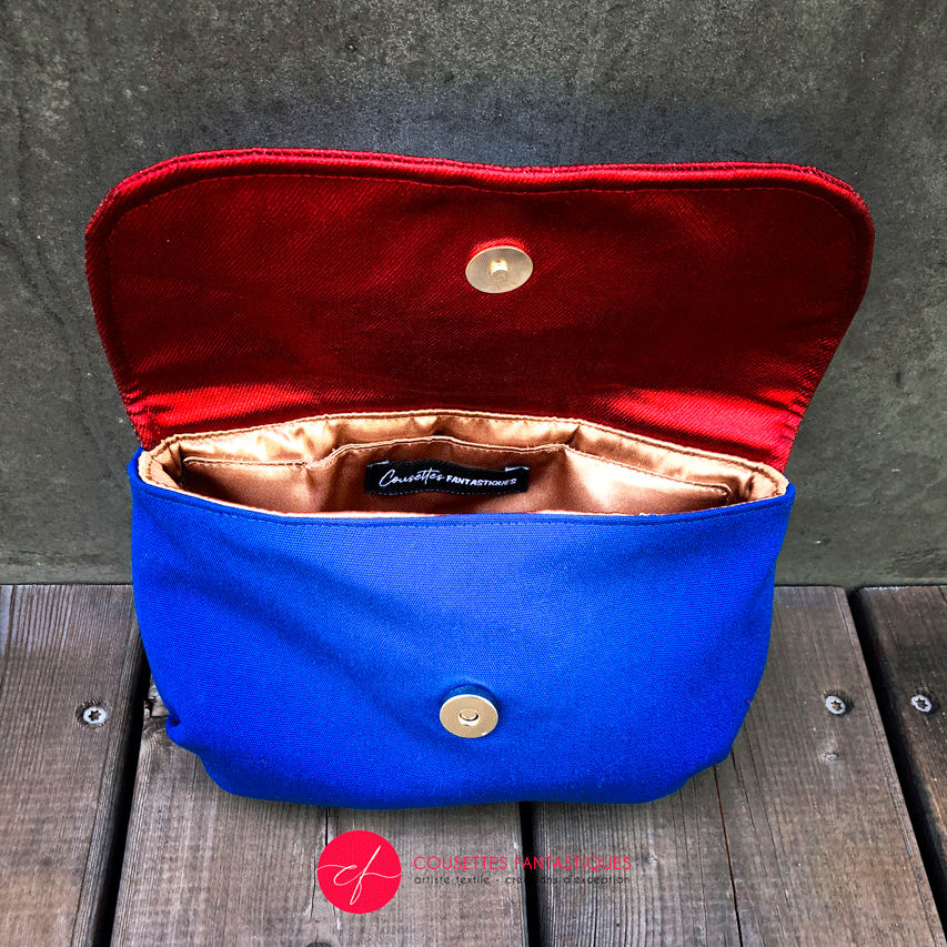 A small flap clutch sewn in blue canvas, metallic red-coated denim, and a glittery avian patterned babywearing wrap, lined with golden satin.