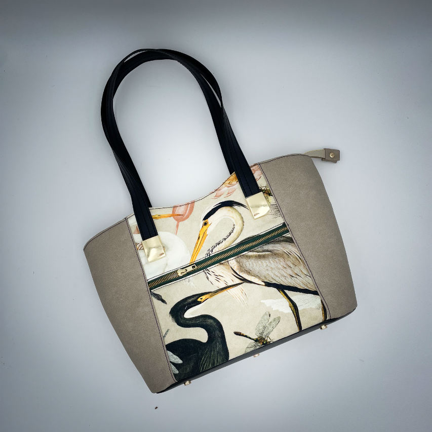 A large zippered shoulder bag, reminiscent of a tote, made from a thick fabric with a wild bird pattern, combined with black and taupe faux leather, and lined with mottled cream linen and coral.