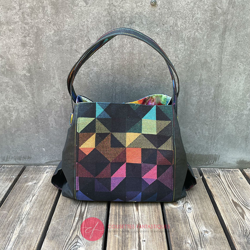 A shoulder bag made from black and rainbow geometric-patterned babywearing scarf and black cork, with a bright rainbow swirl interior.