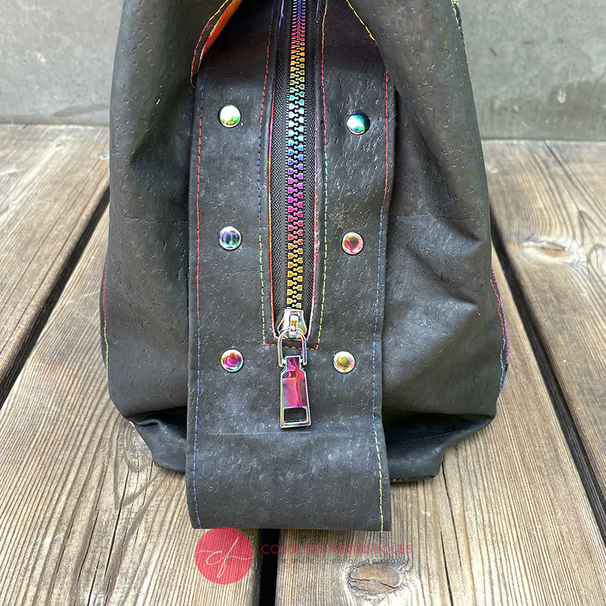 A shoulder bag made from black and rainbow geometric-patterned babywearing scarf and black cork, with a bright rainbow swirl interior.