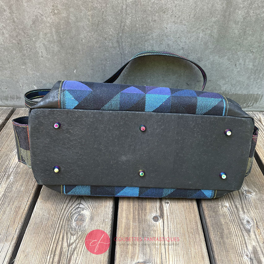 A shoulder bag made from black and rainbow geometric-patterned babywearing scarf and black cork, with a bright rainbow swirl interior.