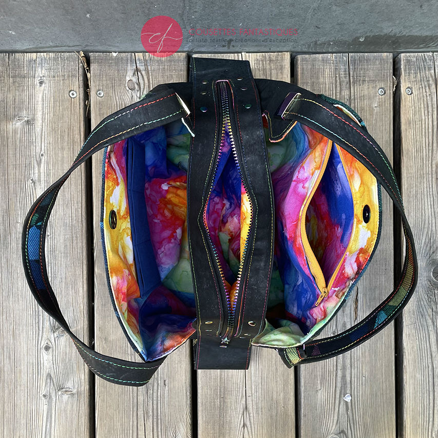 A shoulder bag made from black and rainbow geometric-patterned babywearing scarf and black cork, with a bright rainbow swirl interior.