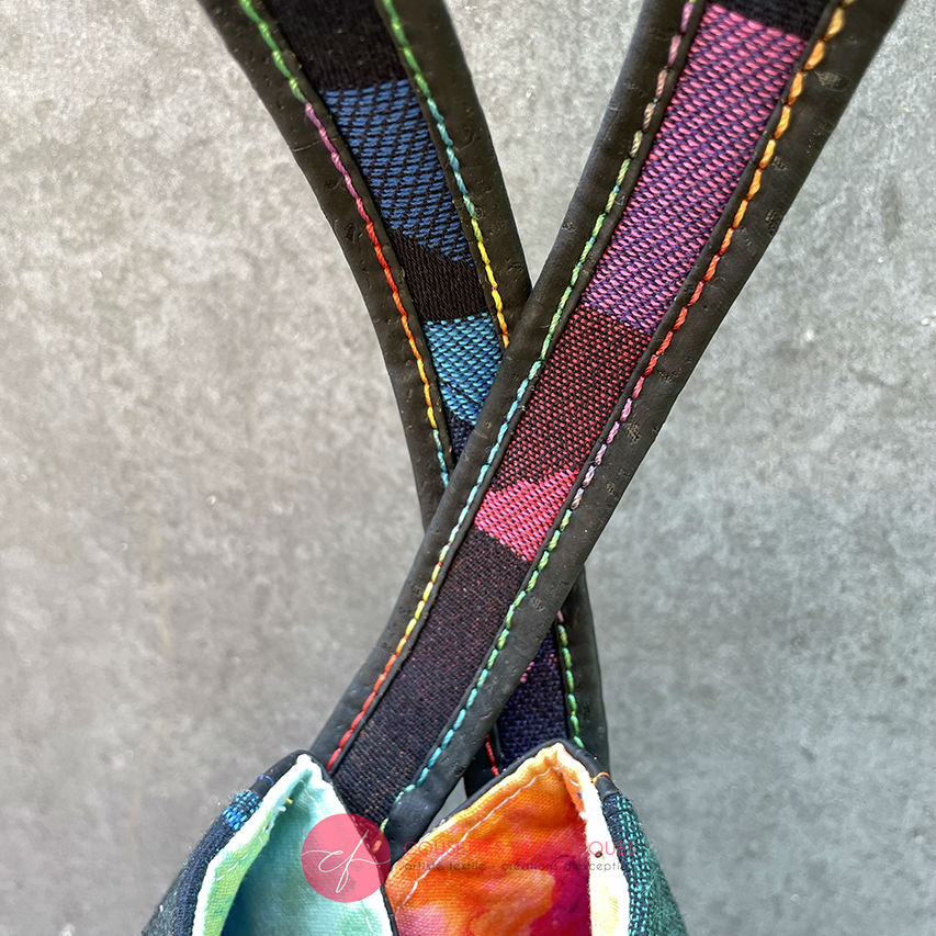 A shoulder bag made from black and rainbow geometric-patterned babywearing scarf and black cork, with a bright rainbow swirl interior.