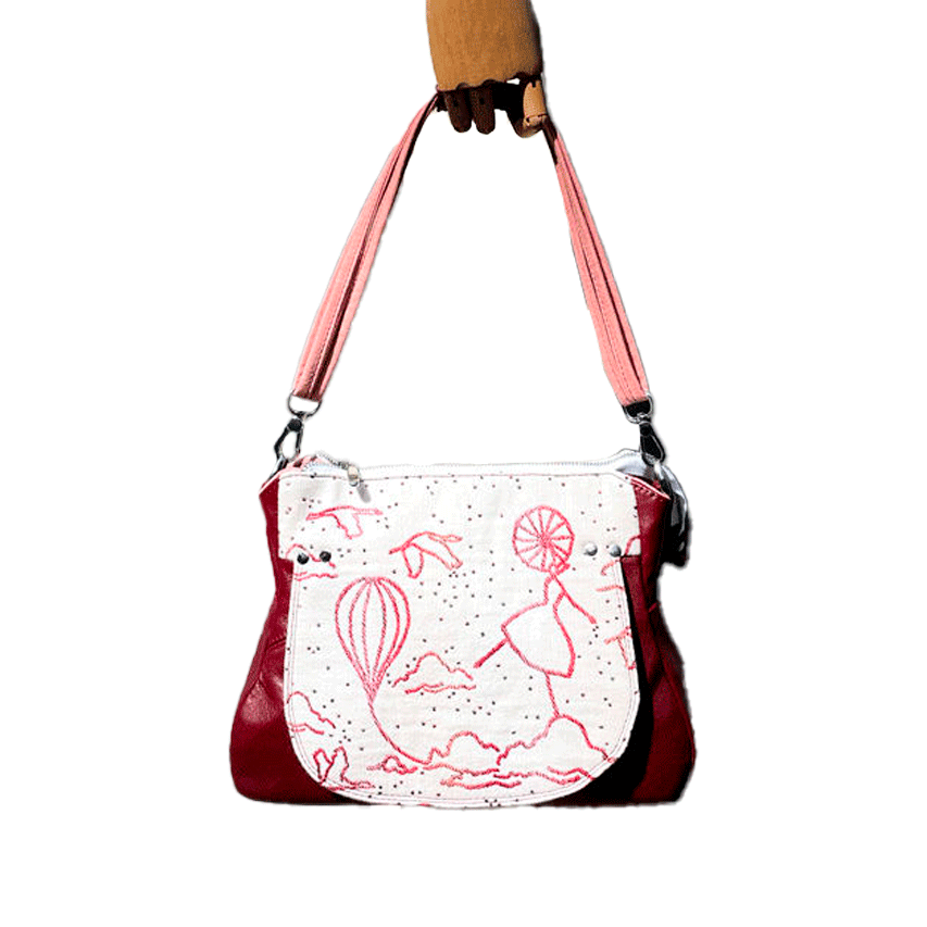 A shoulder bag made with off-white, raspberry, orange, and coral babywearing fabric, featuring a poetic pattern of hot air balloons and dancers, and plum leather.