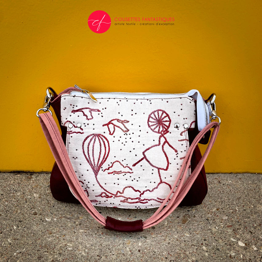 A shoulder bag made with off-white, raspberry, orange, and coral babywearing fabric, featuring a poetic pattern of hot air balloons and dancers, and plum leather.