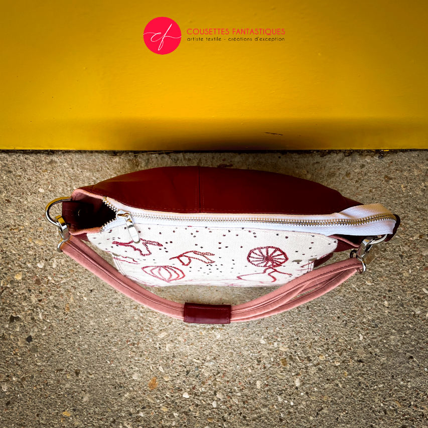 A shoulder bag made with off-white, raspberry, orange, and coral babywearing fabric, featuring a poetic pattern of hot air balloons and dancers, and plum leather.