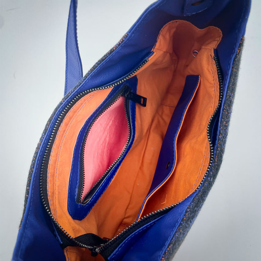 A zippered shoulder bag made from black, blue, pink and orange babywearing wrap scrap, royal blue faux leather, and bright light orange lining.