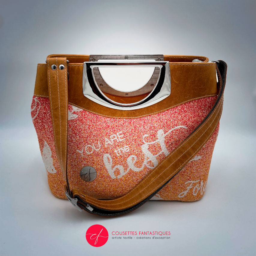 A shoulder bag made of caramel cowhide leather and a gradient-colored babywearing wrap with motivational texts and icons in white.