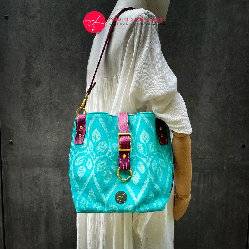 A shoulder bag made of turquoise and ecru babywearing wrap with a leaf pattern, plum synthetic leather, and iridescent mint poplin.
