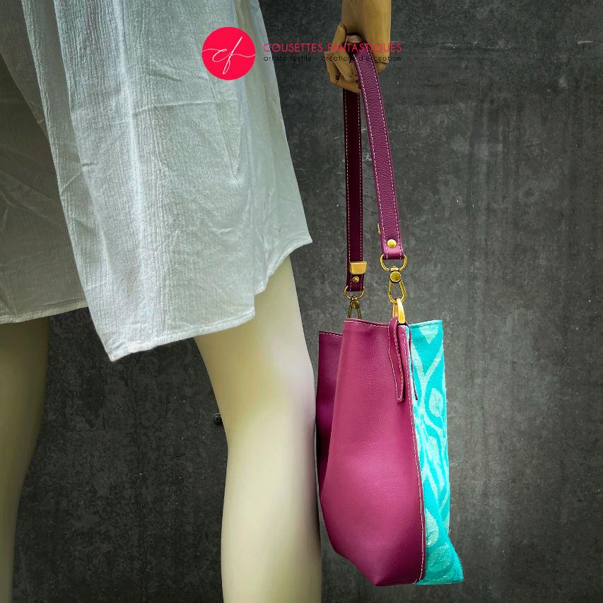A shoulder bag made of turquoise and ecru babywearing wrap with a leaf pattern, plum synthetic leather, and iridescent mint poplin.