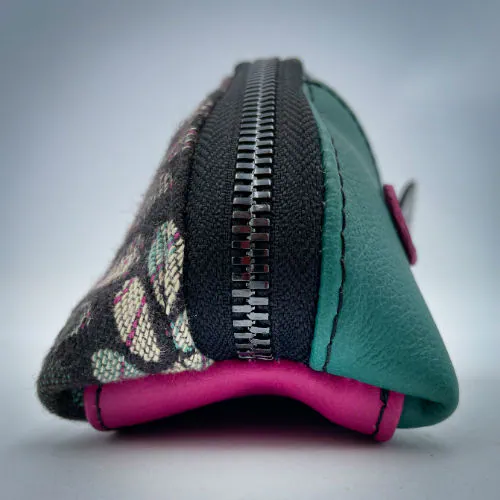 A zippered pouch sewn from sage green leather, black patent leather, fuchsia pink leather, and two scraps of a babywearing wrap with a green, beige, and pink mesh pattern on a black background.