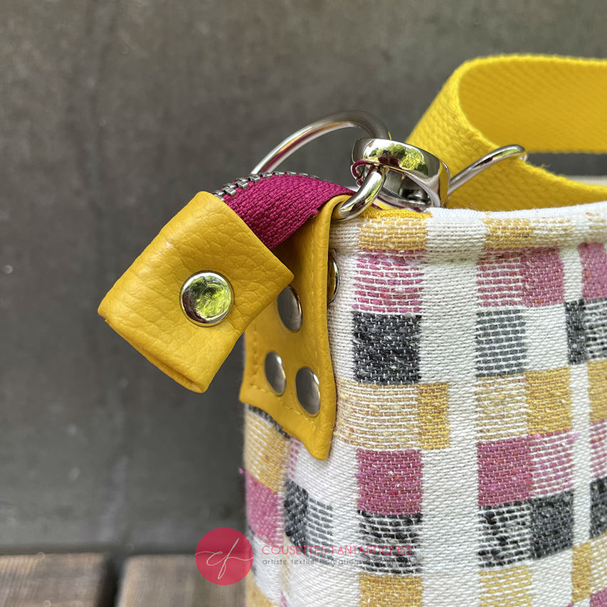 A rectangular messenger bag made from black, white, pink, and yellow babywearing wrap fabric with a geometric pattern, and sunflower yellow faux leather.