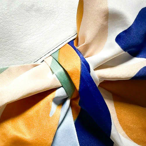 A small zippered pouch sewn in white faux leather and a colorful neck scarf with an abstract and modern flowery design in white, blue, sage, beige and cinammon. Inside is a light sage shiny poplin.