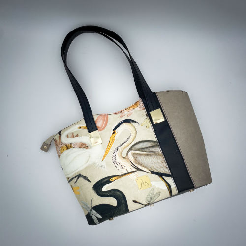 A large zippered shoulder bag, reminiscent of a tote, made from a thick fabric with a wild bird pattern, combined with black and taupe faux leather, and lined with mottled cream linen and coral.